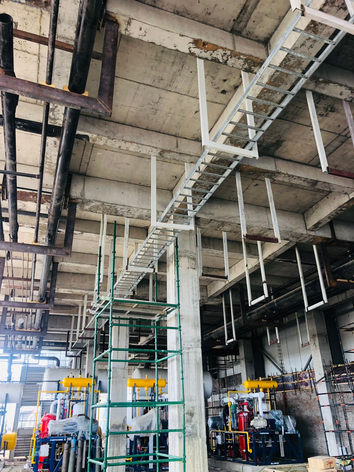 Unfinished industrial space with a complex network of overhead pipes and cable trays, with scaffolding and machinery below.