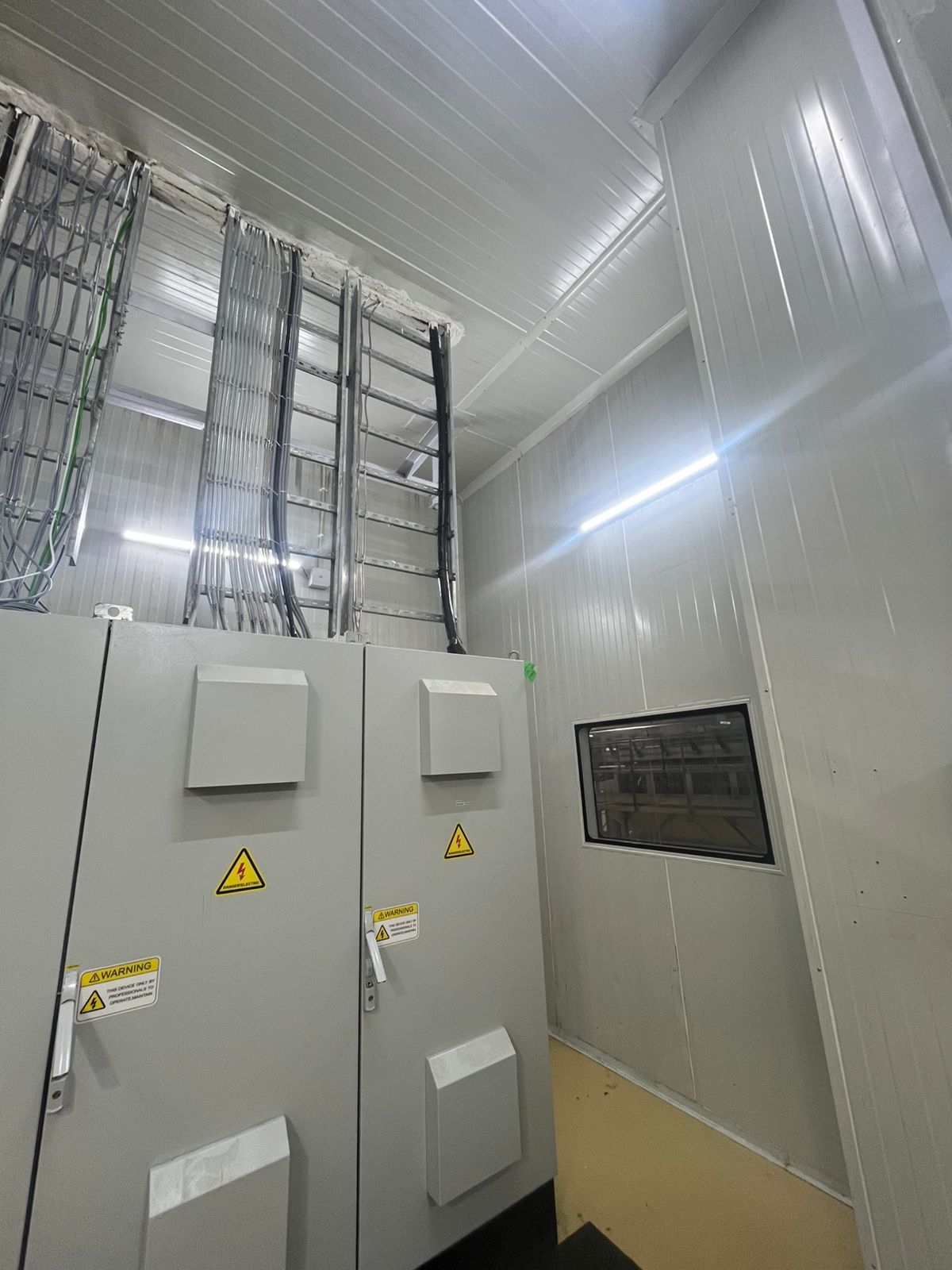 A tall electrical cabinet with warning signs, next to a window and wire racks leading to the ceiling.