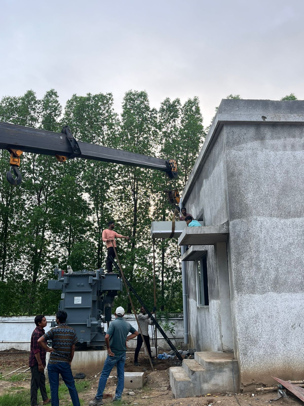 Crane lifting a large electrical transformer onto a concrete pad next to a building under construction.