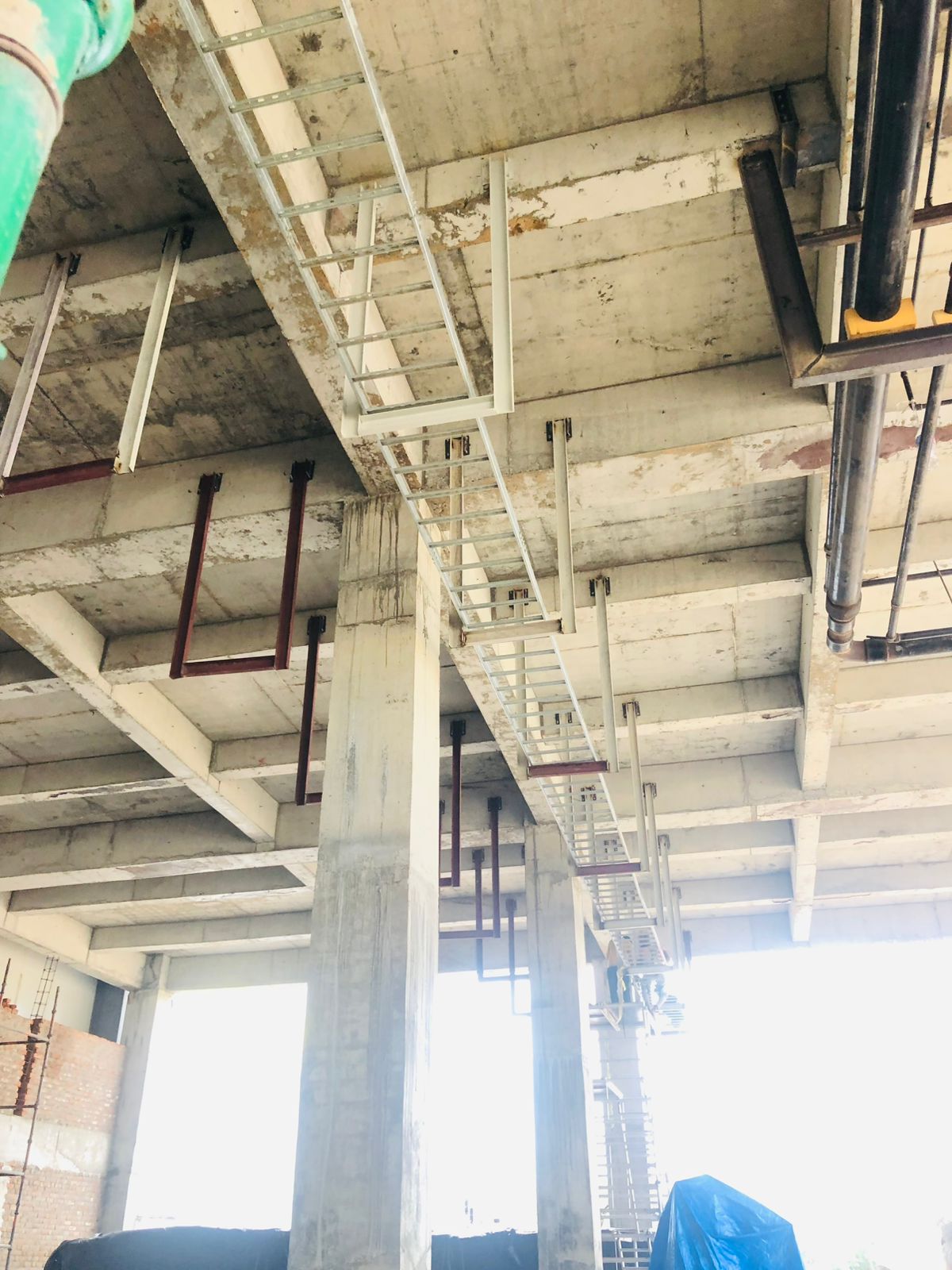 A low-angle view of a building's unfinished concrete ceiling with multiple metal cable trays and pipes suspended below.